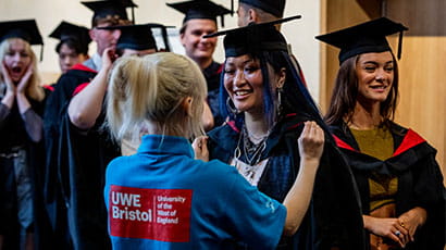 A member of staff in UWE Bristol blue t-shirt helps a student with her gowns in the queue to enter the hall.